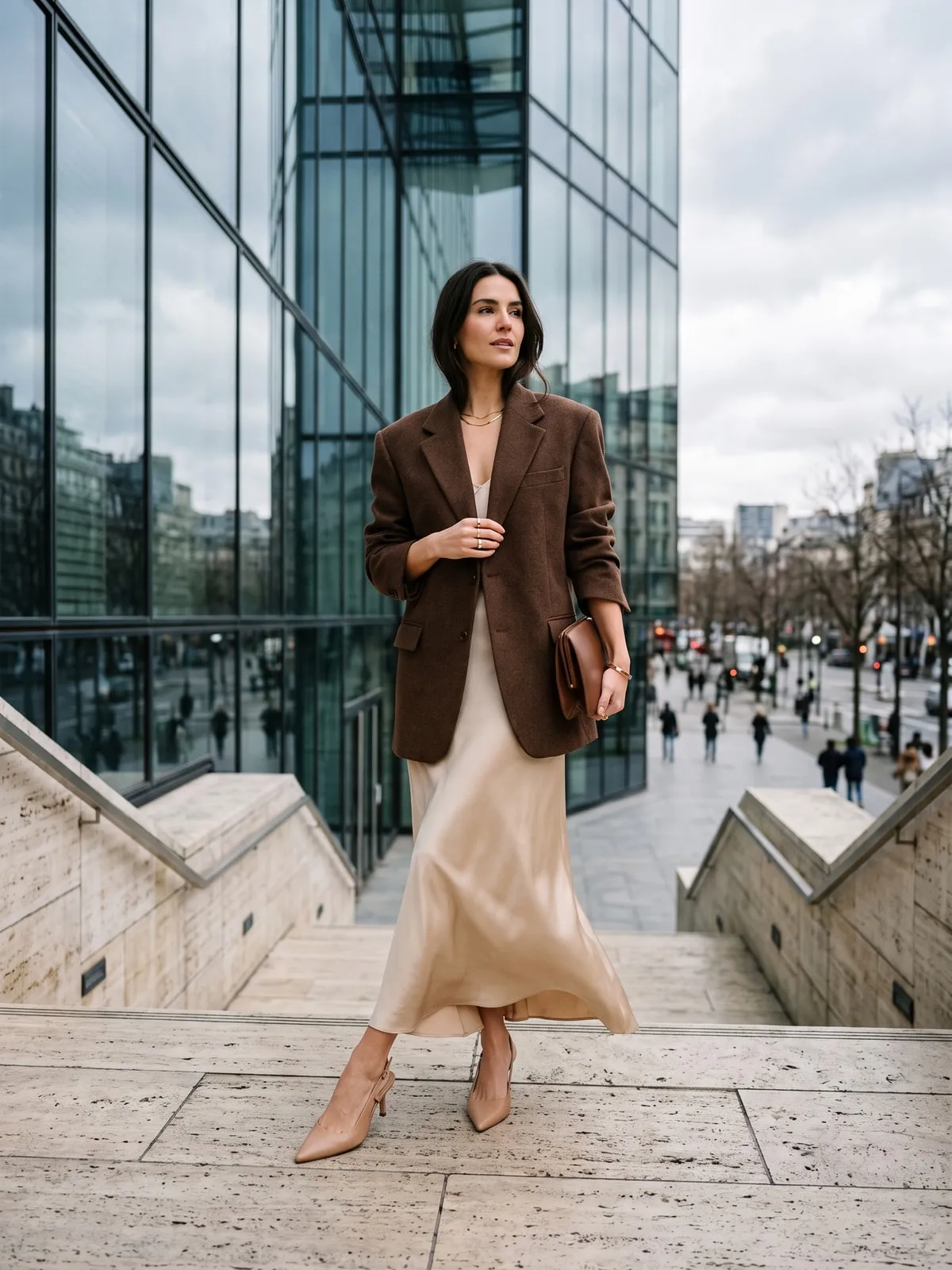 Frau in schokoladenbraunem Oversize-Blazer über einem champagnerfarbenen Satinkleid, nudefarbene Slingpumps, auf einer hellen Steintreppe vor einer Glasfassade